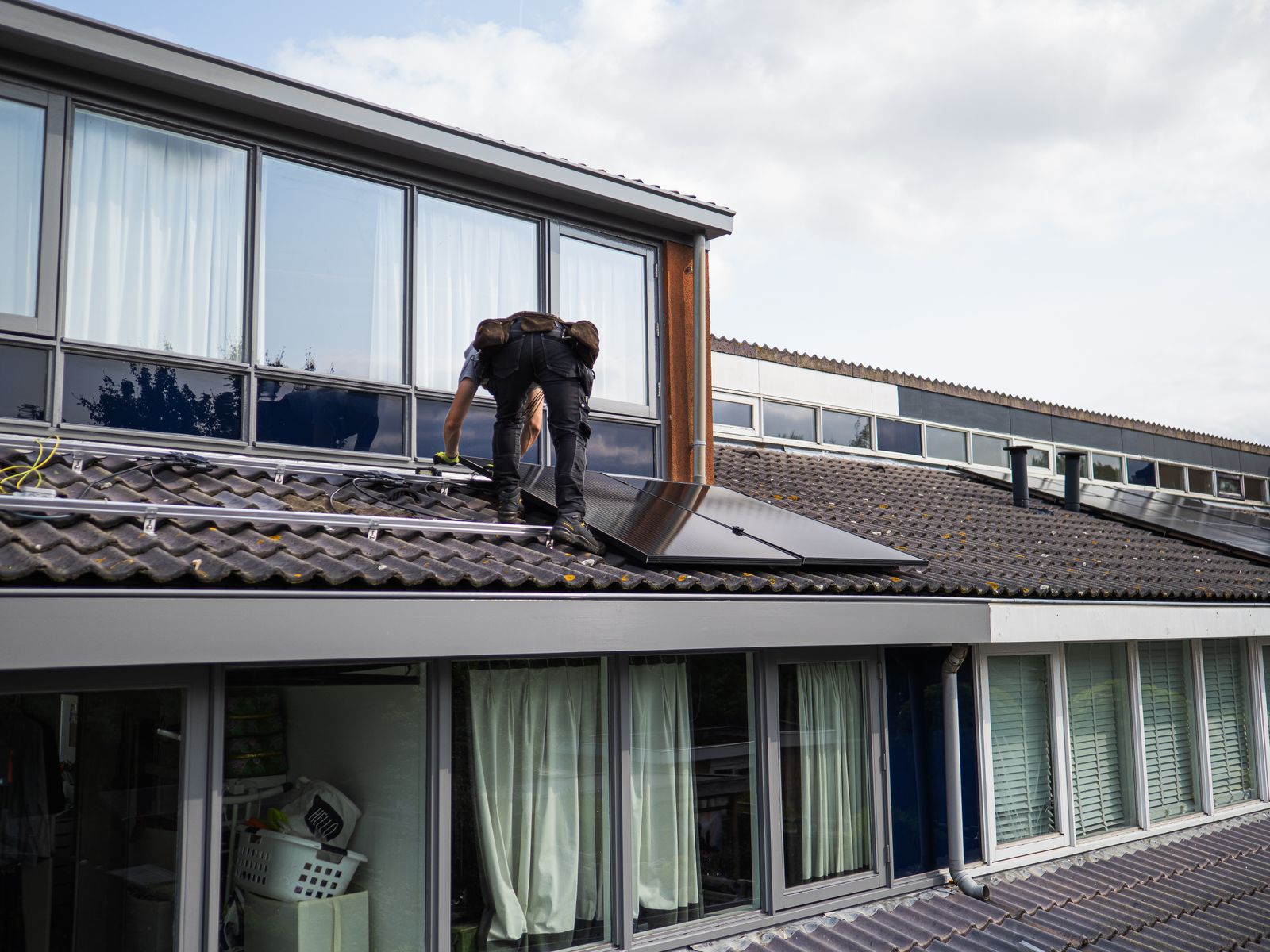 A solar installer kneeling on a tiled roof, guiding a panel into its mounting rail.